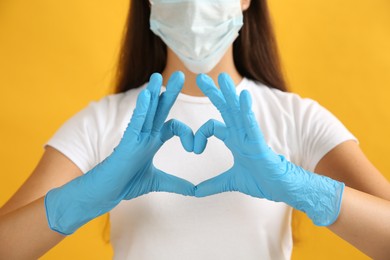 Photo of Woman in protective face mask and medical gloves making heart with hands on yellow background, closeup