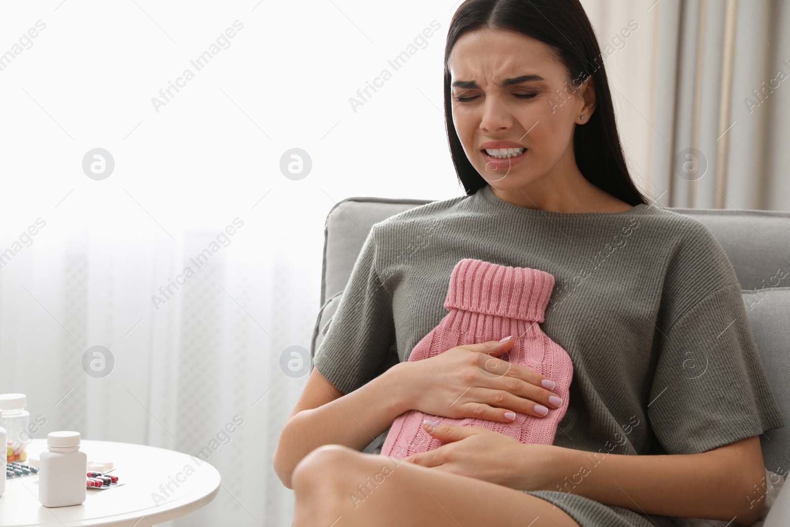 Photo of Woman using hot water bottle to relieve abdominal pain on sofa at home