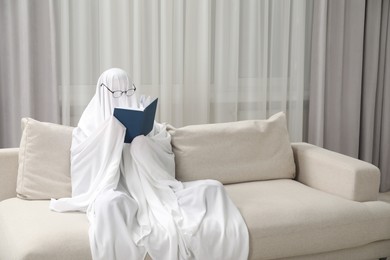 Photo of Creepy ghost. Person covered with white sheet reading book on sofa at home, space for text