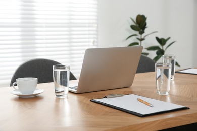 Photo of Modern laptop, glasses of water and clipboard on wooden table in conference room. Interior design