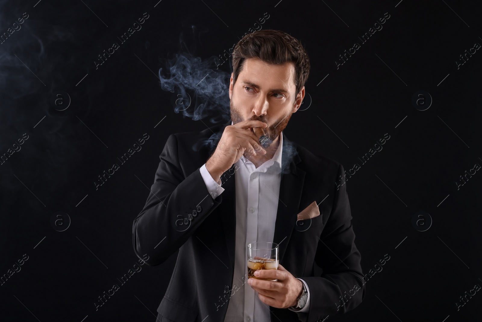 Photo of Handsome man in elegant suit with glass of whiskey smoking cigar on black background