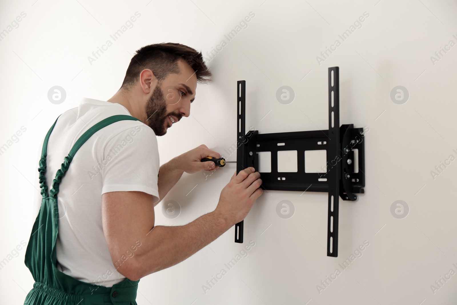 Photo of Professional technician with screwdriver installing TV bracket on wall indoors