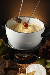 Photo of Dipping pieces of bread and grape into fondue pot with melted cheese at table with snacks, closeup