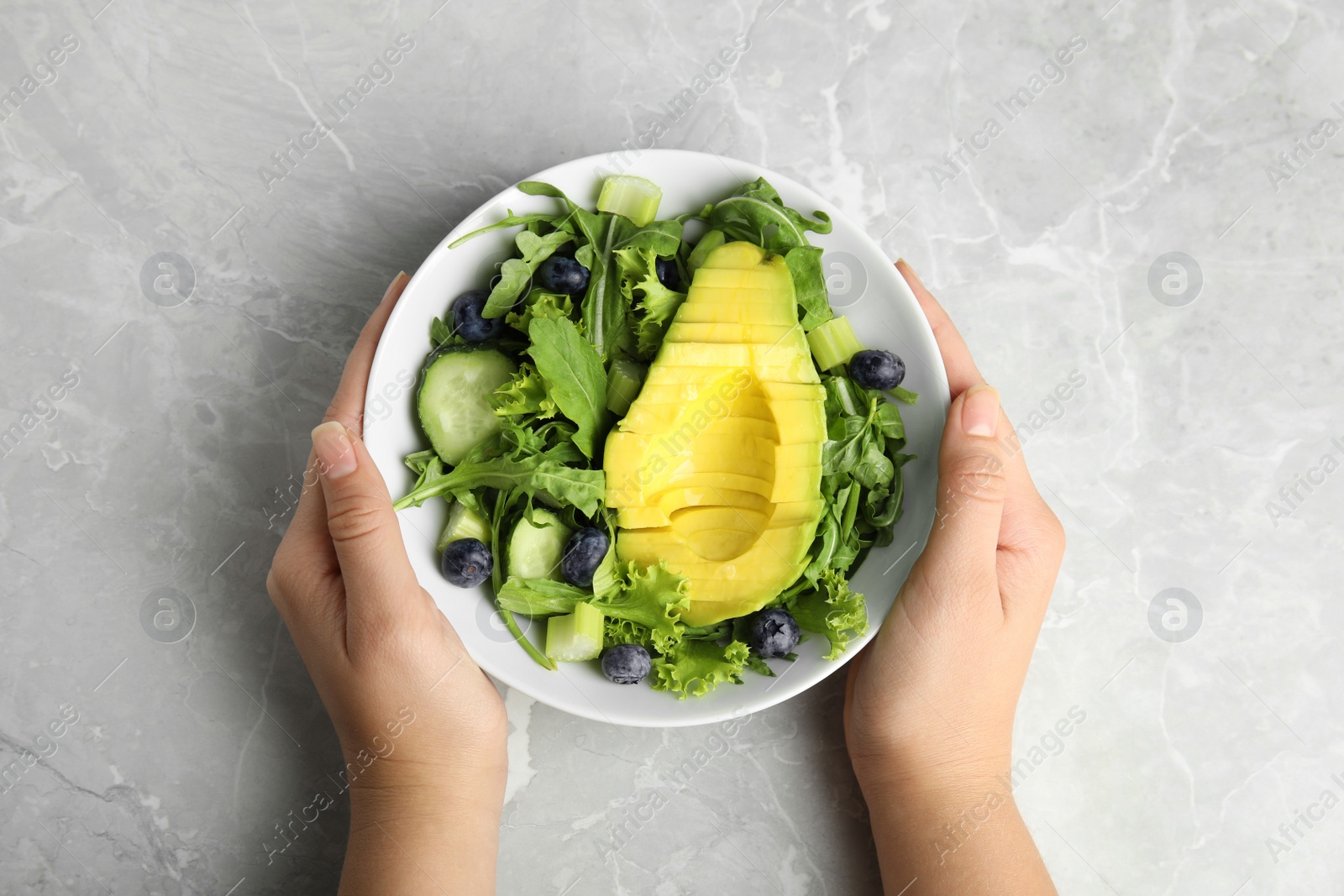 Photo of Woman holding bowl of avocado salad with blueberries over grey marble table, top view