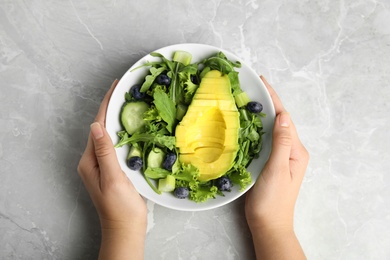 Woman holding bowl of avocado salad with blueberries over grey marble table, top view