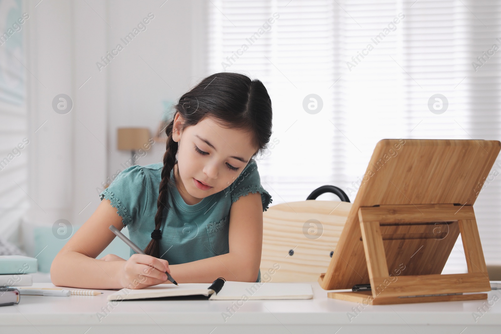 Photo of Little girl doing homework with tablet at table in room