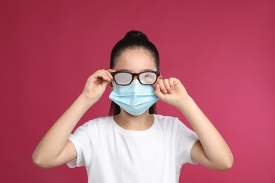 Little girl wiping foggy glasses caused by wearing medical face mask on pink background. Protective measure during coronavirus pandemic