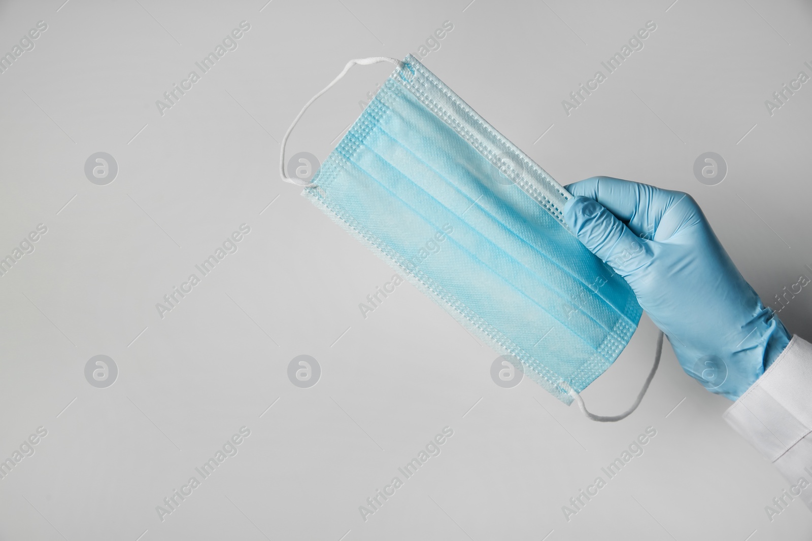 Photo of Doctor in latex gloves holding disposable face mask on light background, closeup with space for text. Protective measures during coronavirus quarantine