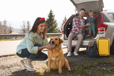 Photo of Happy woman petting dog, father and his daughter sitting in car trunk near road. Family traveling with pet