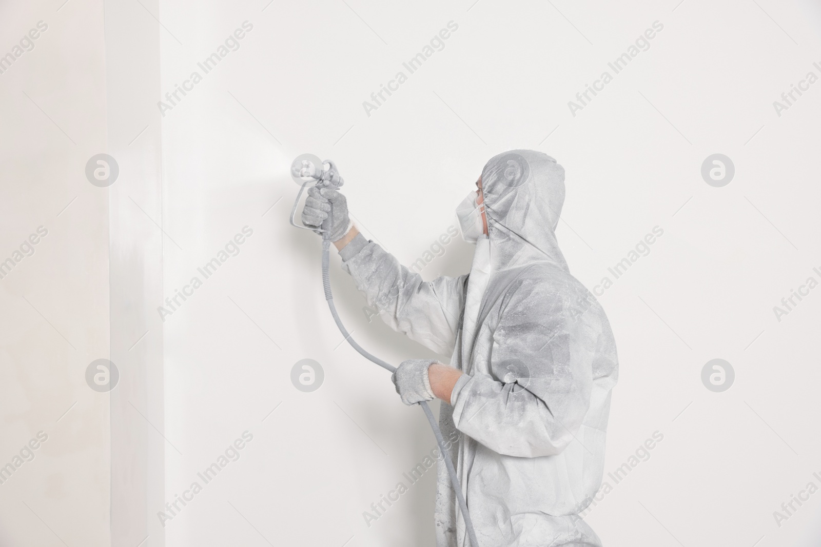 Photo of Decorator in protective overalls painting wall with spray gun indoors