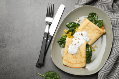 Delicious thin pancakes with spinach and sour cream on grey table, flat lay