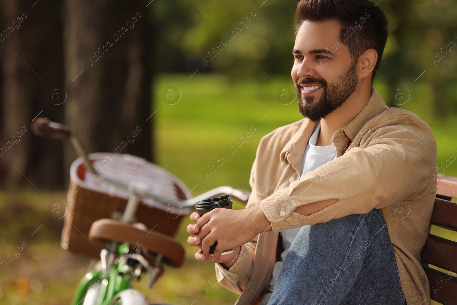 Photo of Young man sitting on bench and holding cup of coffee in park, space for text