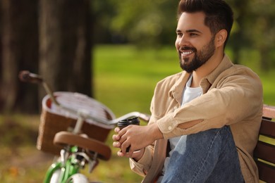 Photo of Young man sitting on bench and holding cup of coffee in park, space for text