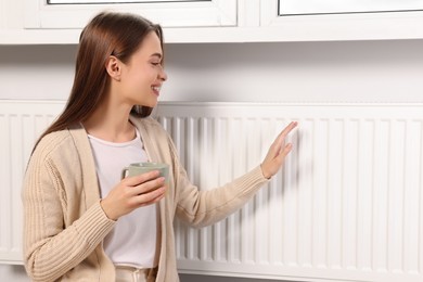 Photo of Woman holding cup with hot drink and warming hand on heating radiator indoors