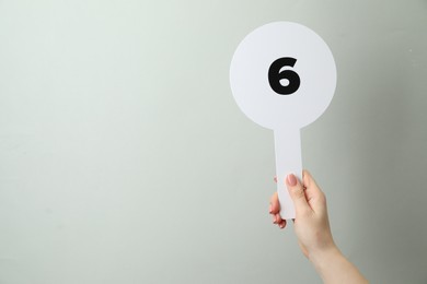 Photo of Woman holding auction paddle with number 6 on light grey background, closeup. Space for text