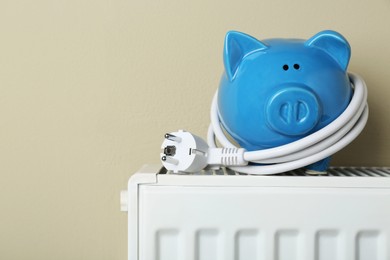 Photo of Piggy bank with plug on heating radiator against beige background, space for text