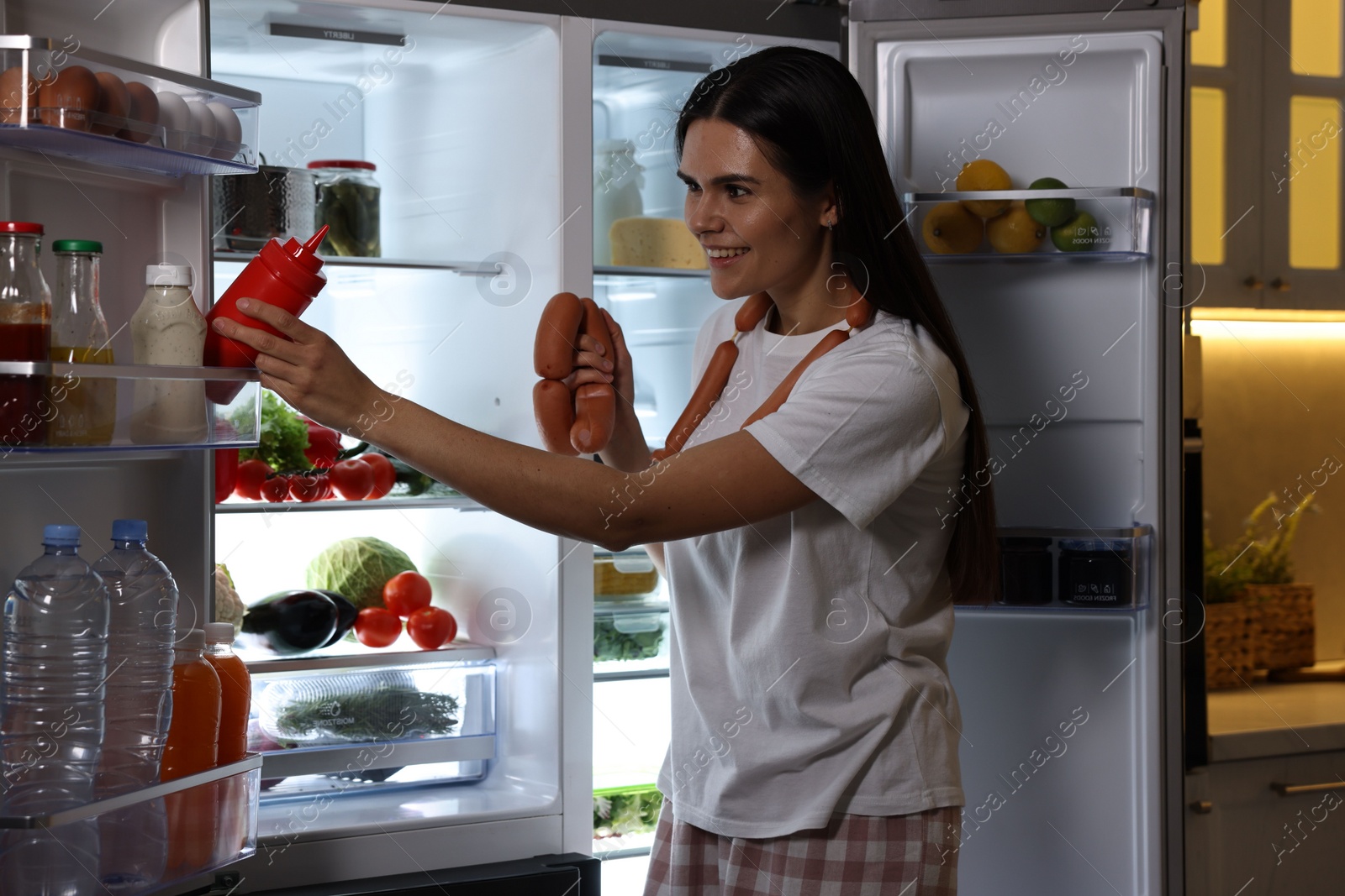 Photo of Young woman taking ketchup and sausages out of refrigerator in kitchen at night