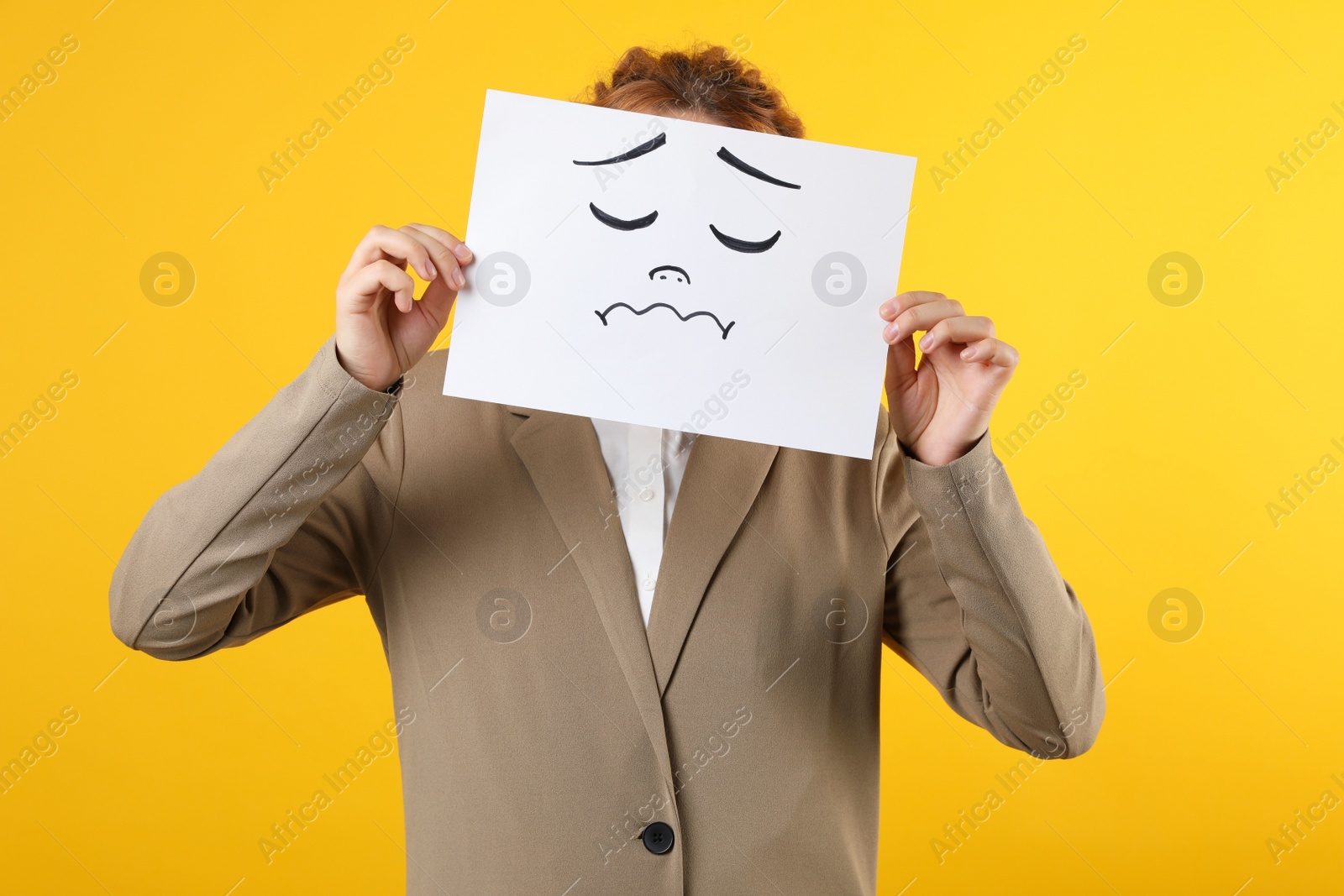 Photo of Woman hiding behind sheet of paper with sad face on yellow background
