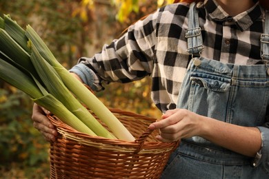 Photo of Woman holding wicker basket with fresh raw leeks outdoors, closeup