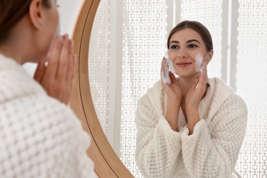 Photo of Young woman applying cleansing foam onto her face near mirror in bathroom