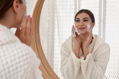 Young woman applying cleansing foam onto her face near mirror in bathroom