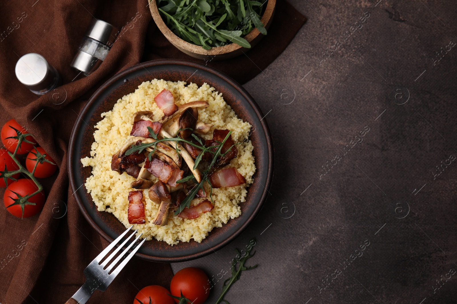 Photo of Tasty couscous with mushrooms and bacon in bowl served on brown table, flat lay. Space for text
