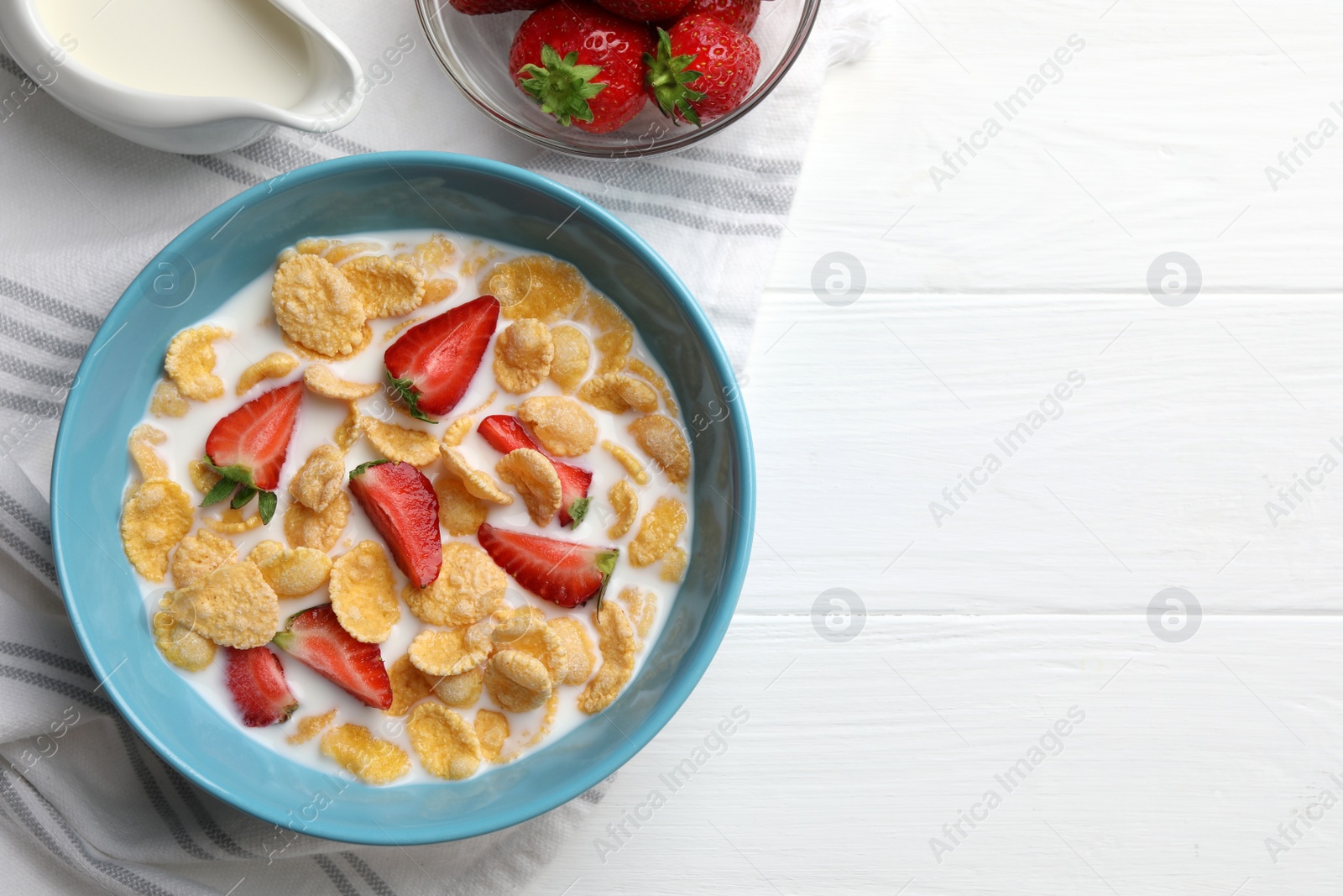 Photo of Bowl of tasty crispy corn flakes with milk and strawberries on white wooden table, flat lay. Space for text