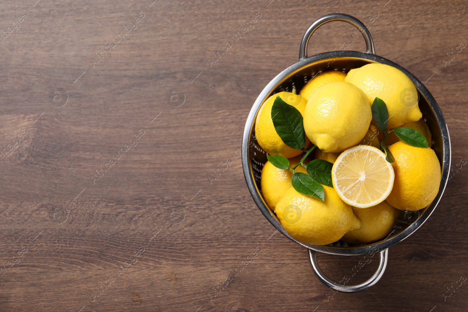 Photo of Fresh lemons and green leaves in colander on wooden table, top view. Space for text