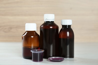 Photo of Bottles of cough syrup, dosing spoon and measuring cup on white wooden table