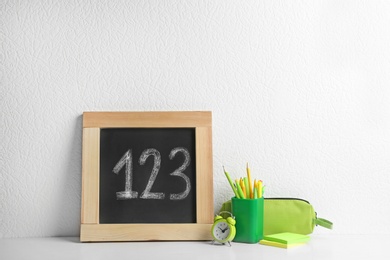Photo of Small chalkboard with numbers and different school stationery on wooden table near white wall