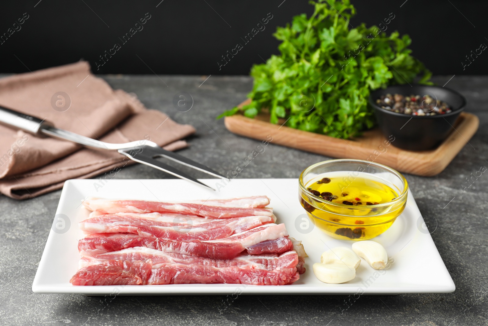 Photo of Pieces of raw pork belly, oil, parsley and garlic on grey textured table