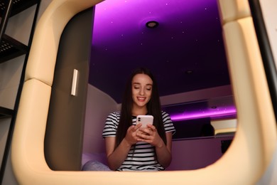 Photo of Happy young woman using smartphone in capsule of pod hostel