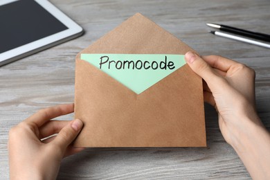 Woman holding envelope and card with words Promo Code at wooden table, closeup