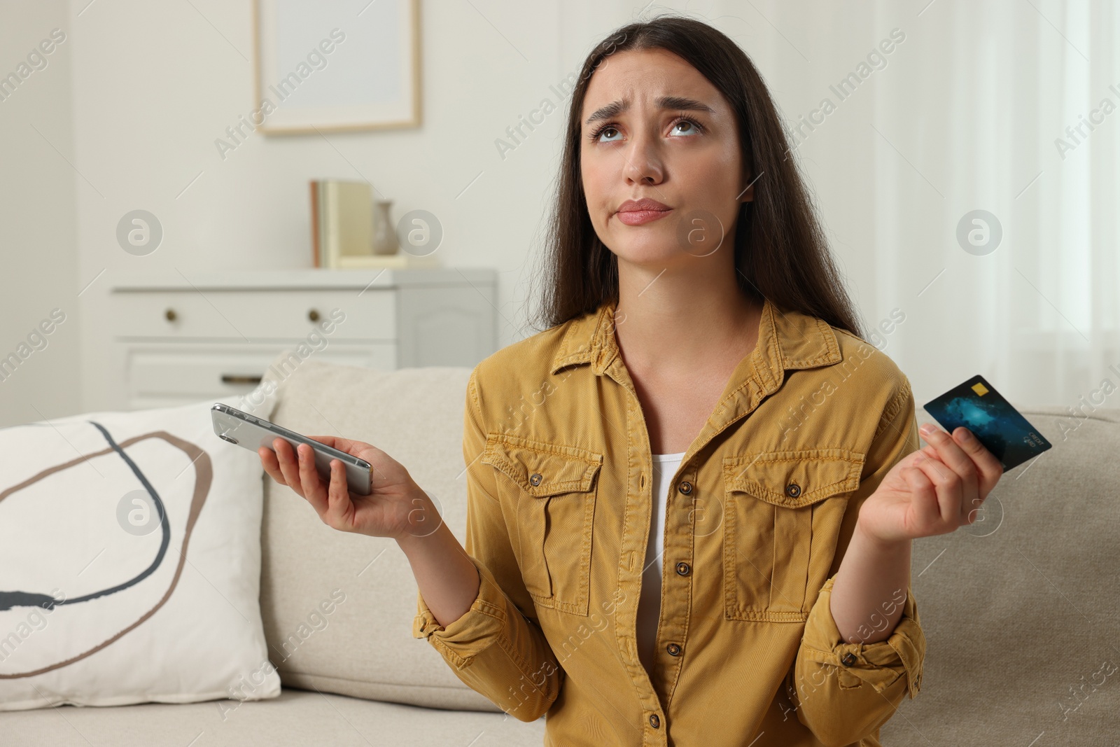 Photo of Confused woman with credit card and smartphone at home. Be careful - fraud