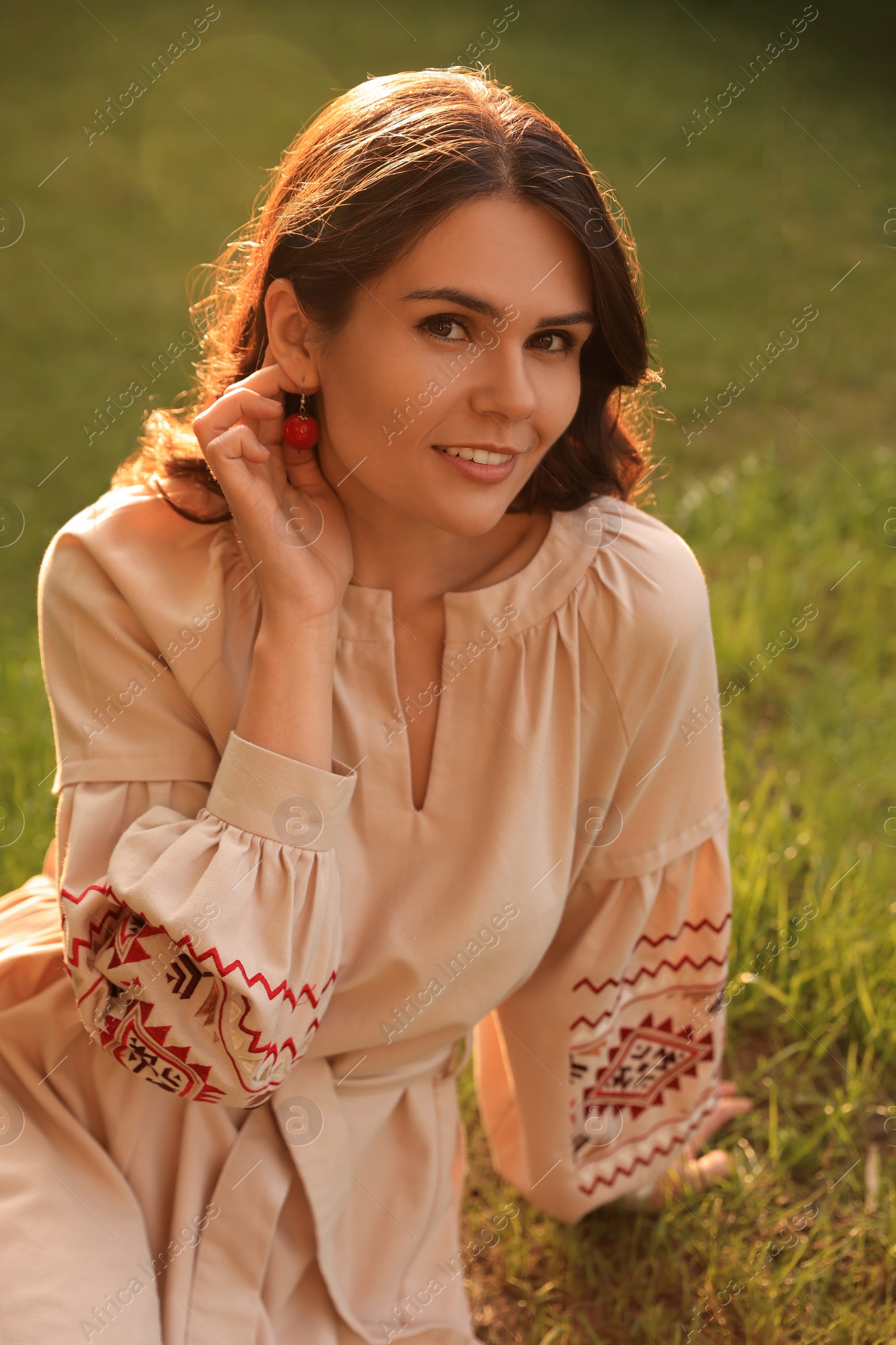 Photo of Beautiful woman in embroidered dress sitting on green grass outdoors. Ukrainian national clothes