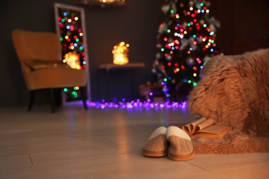 Photo of Soft slippers with fluffy blanket and Christmas tree in dark room