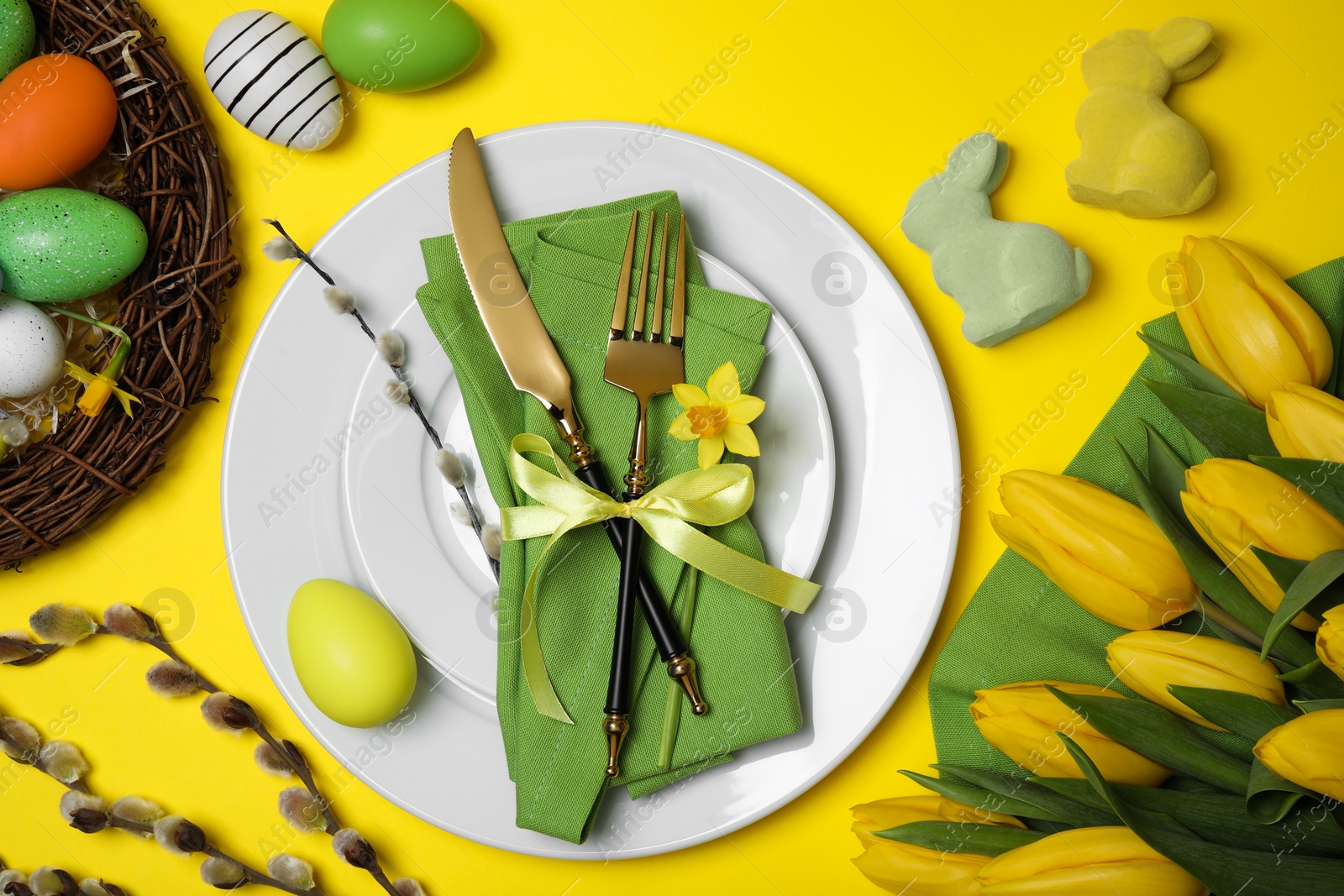 Photo of Festive table setting with painted eggs and tulips on yellow background, flat lay. Easter celebration