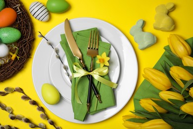 Photo of Festive table setting with painted eggs and tulips on yellow background, flat lay. Easter celebration