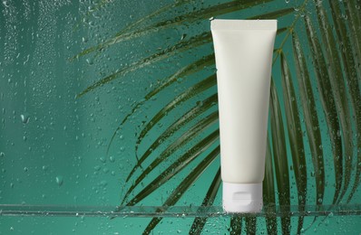 Tube with moisturizing cream and palm leaf on green background, view through wet glass