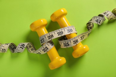 Photo of Measuring tape and dumbbells on light green background, flat lay. Weight control concept