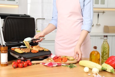 Woman cooking different products with electric grill at wooden table in kitchen, closeup