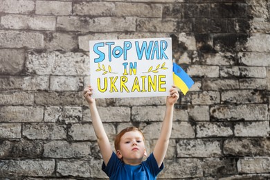 Photo of Sad boy holding poster Stop War In Ukraine and national flag against brick wall outdoors