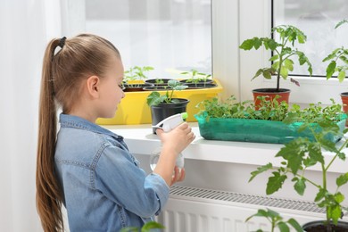 Photo of Cute little girl spraying seedlings in plastic container on windowsill indoors