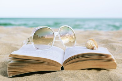 Photo of Beautiful sunglasses, book and shell on sand near sea, closeup