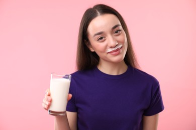 Cute woman with milk mustache holding glass of tasty dairy drink on pink background