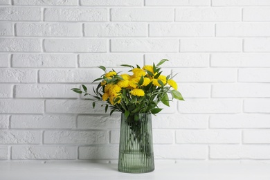 Photo of Bouquet of beautiful yellow flowers in glass vase on white wooden table near brick wall