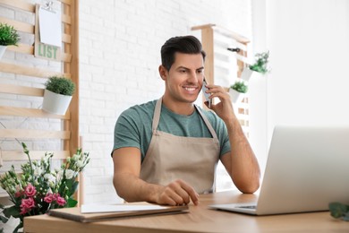 Photo of Florist talking on smartphone near laptop in workshop