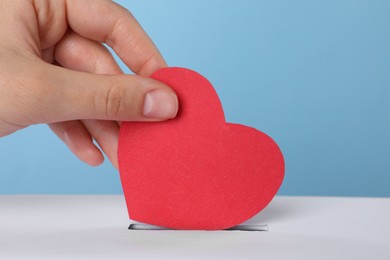 Woman putting red heart into slot of donation box against light blue background, closeup