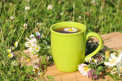 Green cup with tea, different wildflowers and herbs on wooden board in meadow