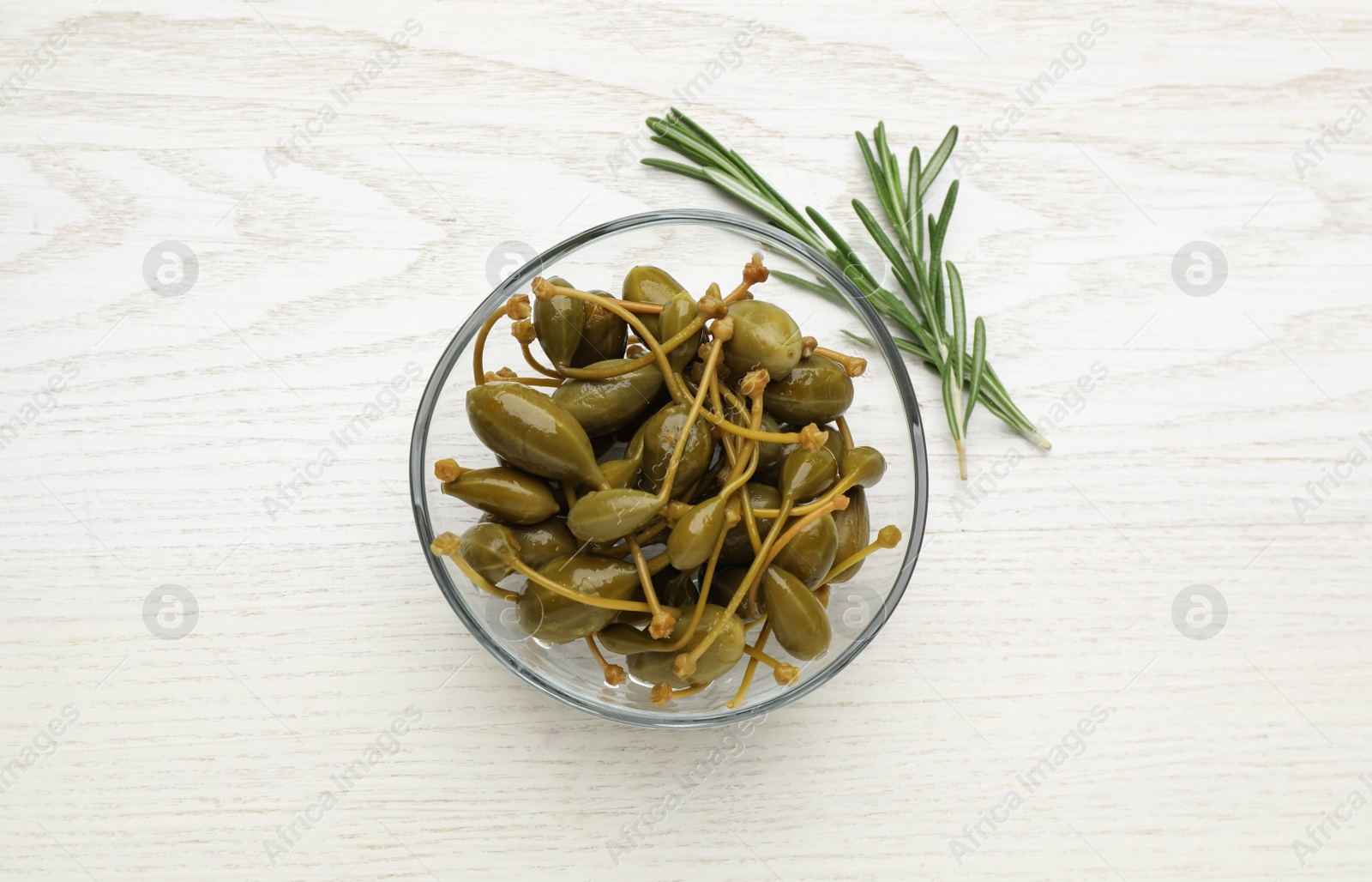 Photo of Tasty capers in bowl and rosemary on white wooden table, top view